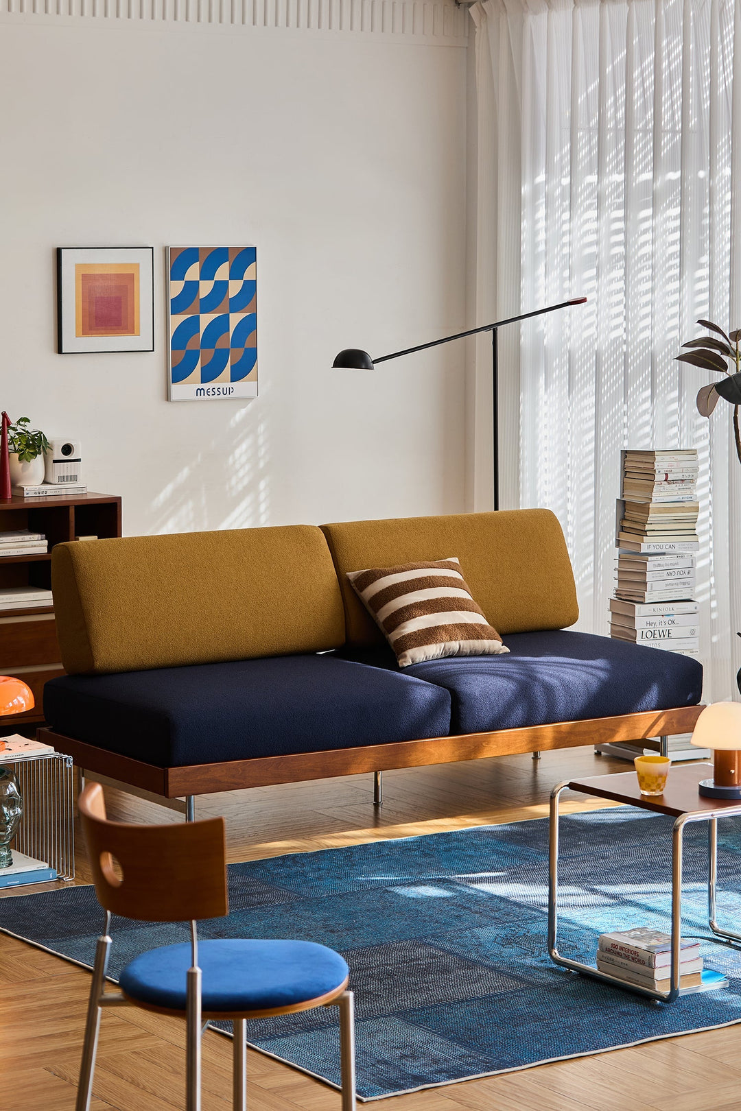 Bauhaus-style two-seater sofa with mustard and navy cushions, supported by a wooden frame and steel legs, adding modern flair to a sunlit living room.