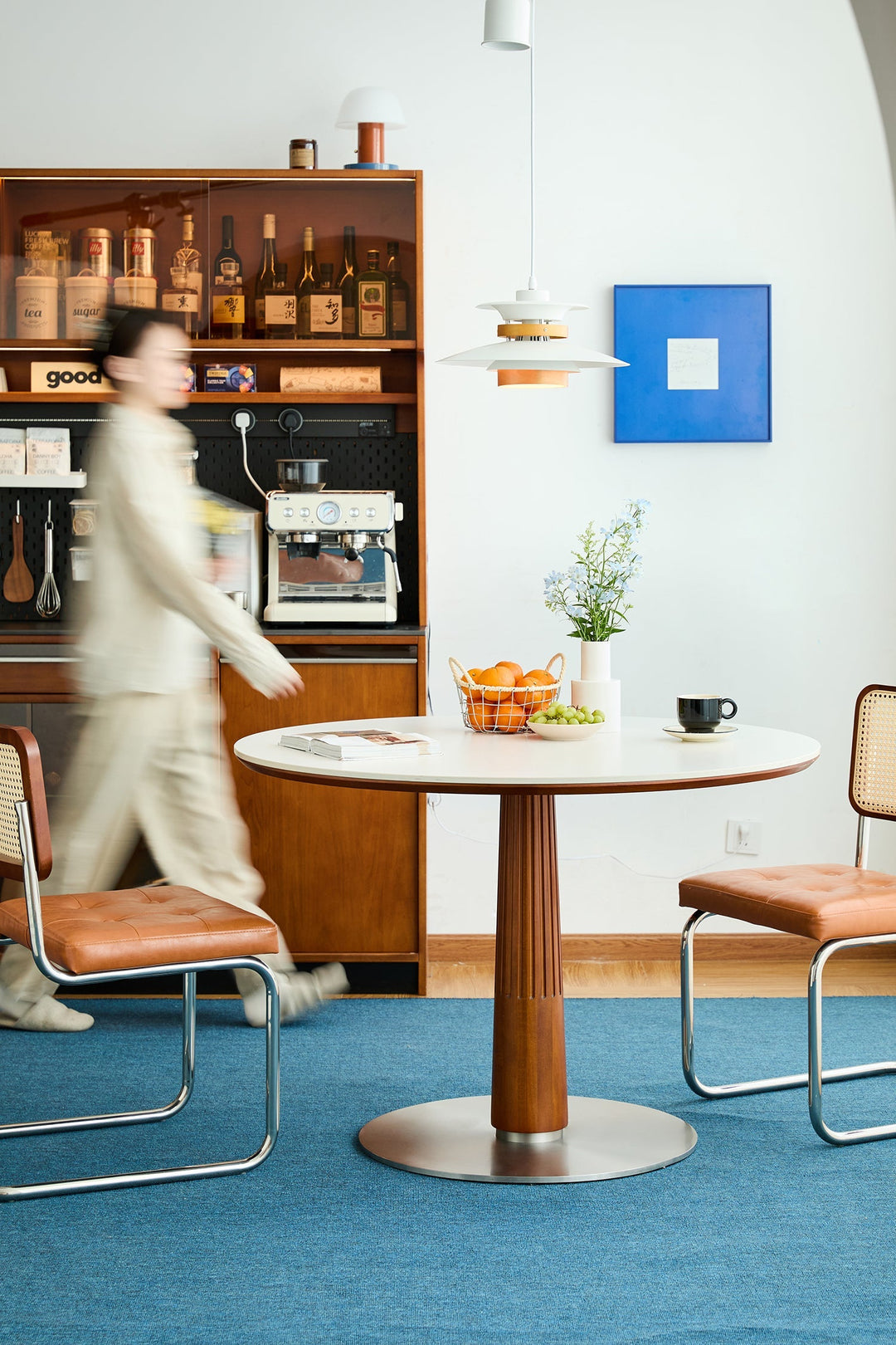 Round Bauhaus-inspired dining table with fluted wood pedestal base and two leather-seated cantilever chairs, styled in a cozy café corner with coffee bar and pendant light.