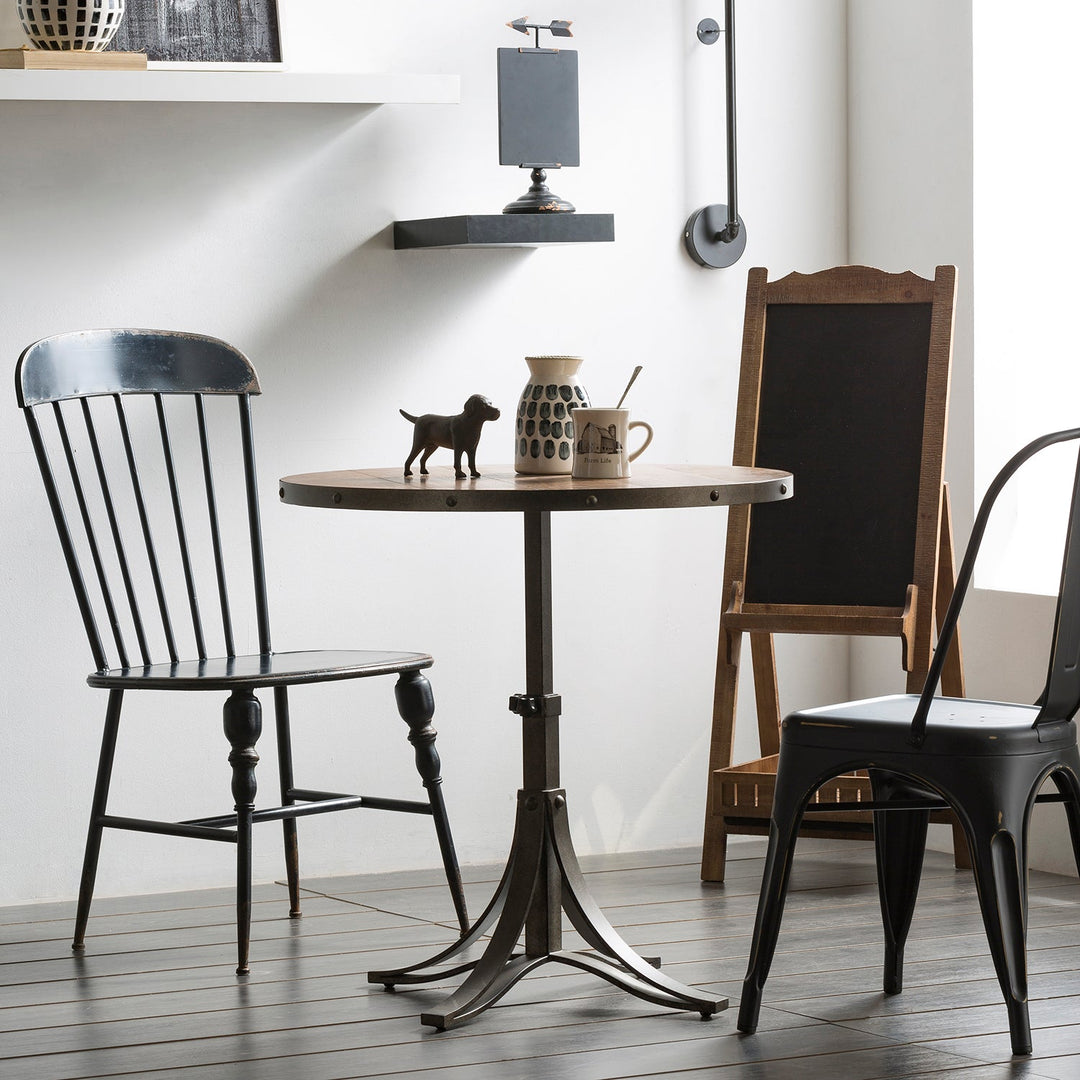 Compact industrial-style dining set with a round table and two mismatched black metal chairs. The setting includes minimalist decor elements like a ceramic jug, a small animal figure, and a chalkboard, creating a cozy, eclectic ambiance.