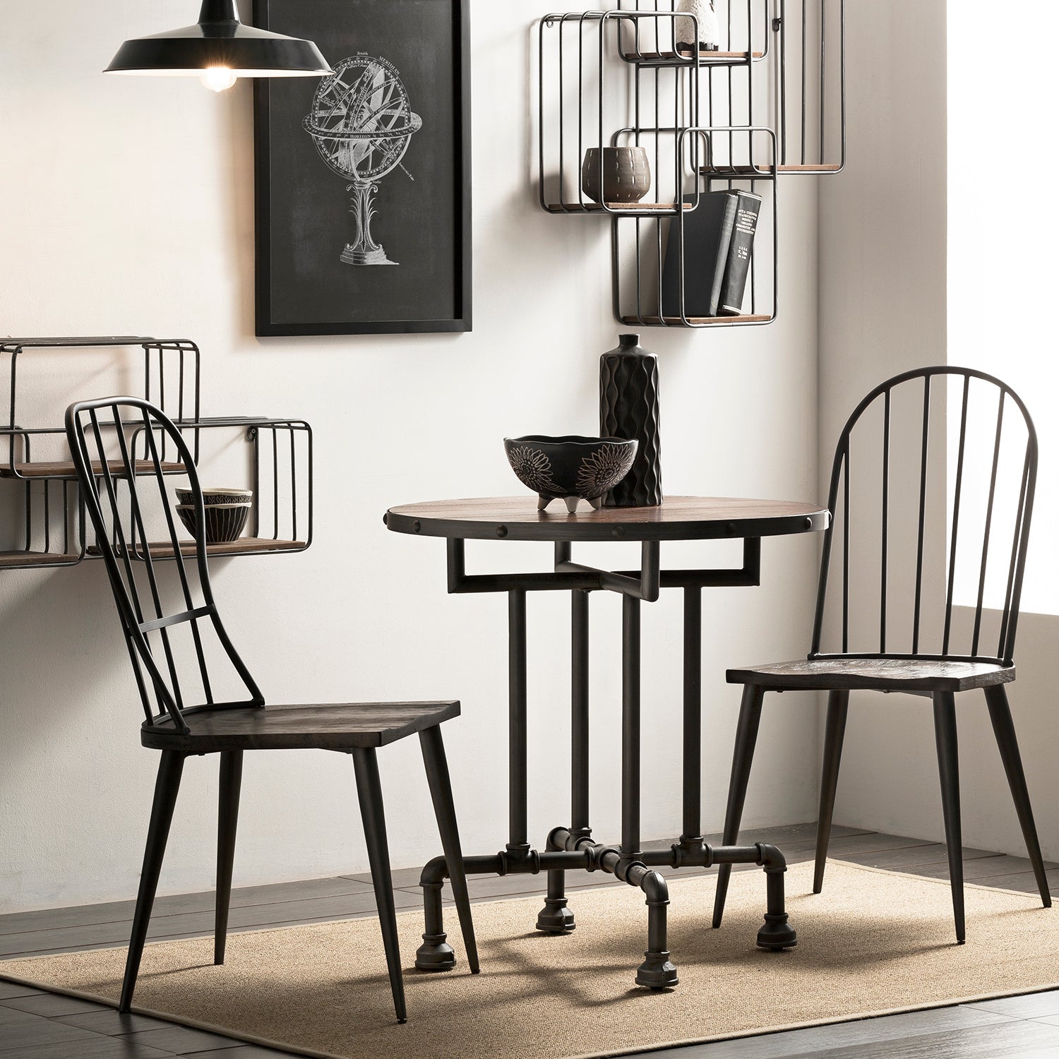 Industrial-style dining set featuring a round table with black metal pipe legs and two matching black chairs, set against a minimalistic background with wire-frame wall shelves and modern decor.
